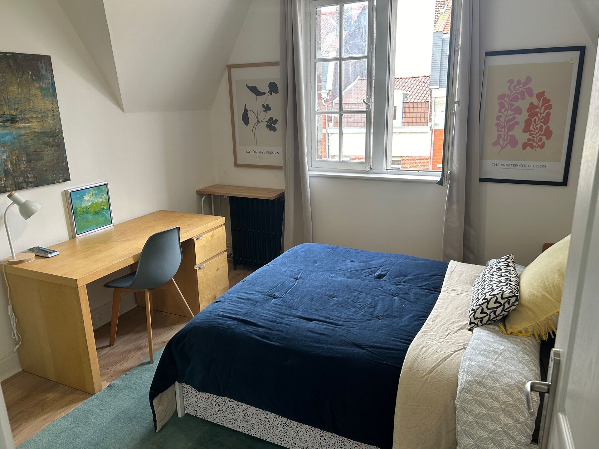 A bedroom with a bed, a desk, and a chair. There is a window with curtains, and artwork on the walls.