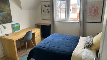A bedroom with a bed, a desk, and a chair. There is a window with curtains, and artwork on the walls.