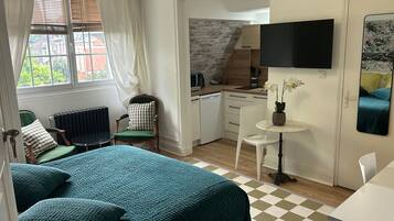 A compact bedroom with a bed, a small sitting area with a chair, a table with a vase, and a kitchen area with a TV.