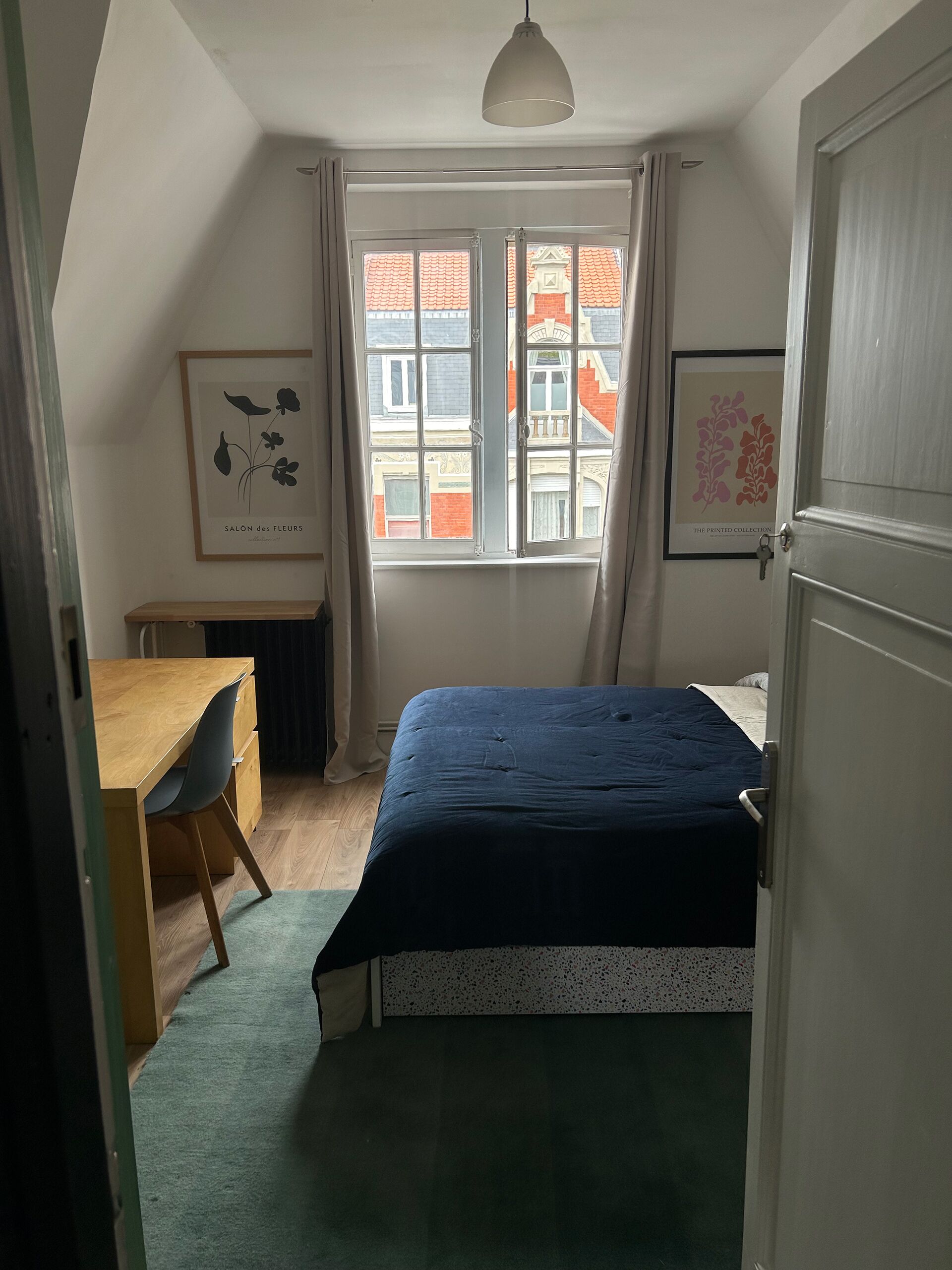 A bedroom with a bed, a desk, and a chair. There is a window with curtains, and two framed pictures on the wall.