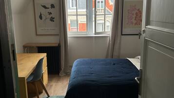 A bedroom with a bed, a desk, and a chair. There is a window with curtains, and two framed pictures on the wall.
