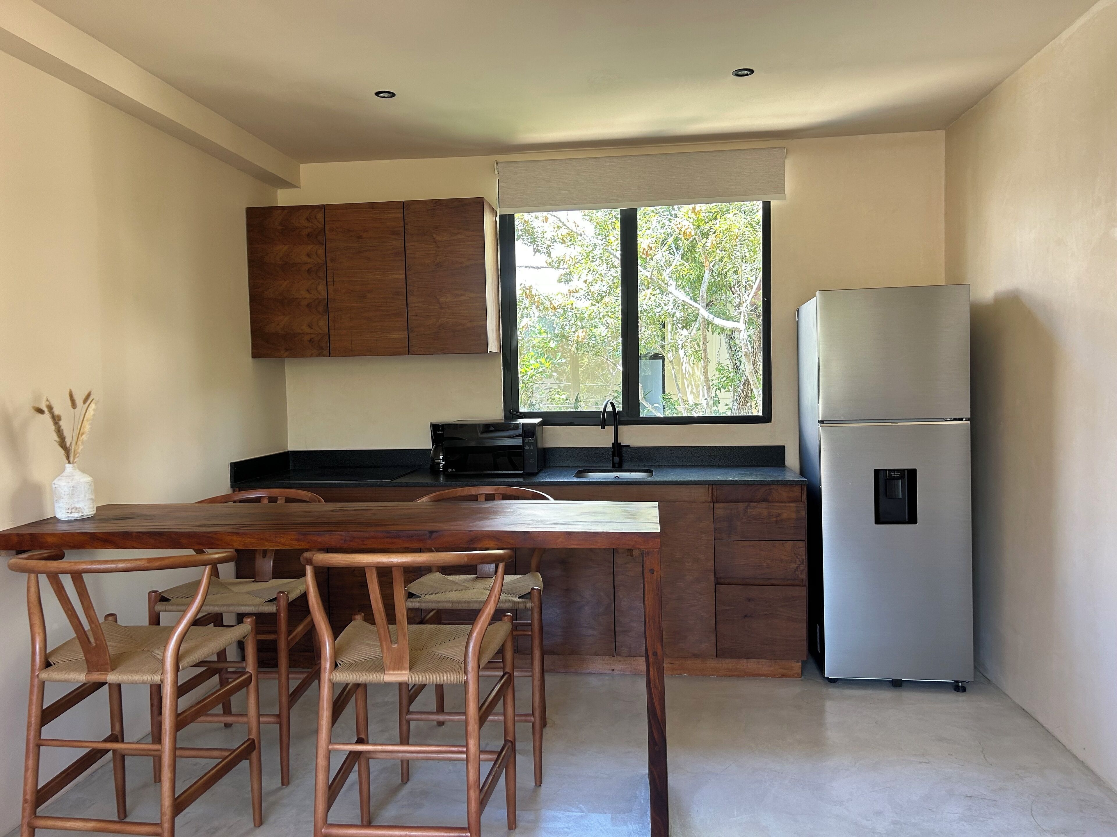 Une cuisine moderne avec des meubles en bois, un réfrigérateur en acier inoxydable, une table à manger avec des chaises et une fenêtre donnant sur les arbres.