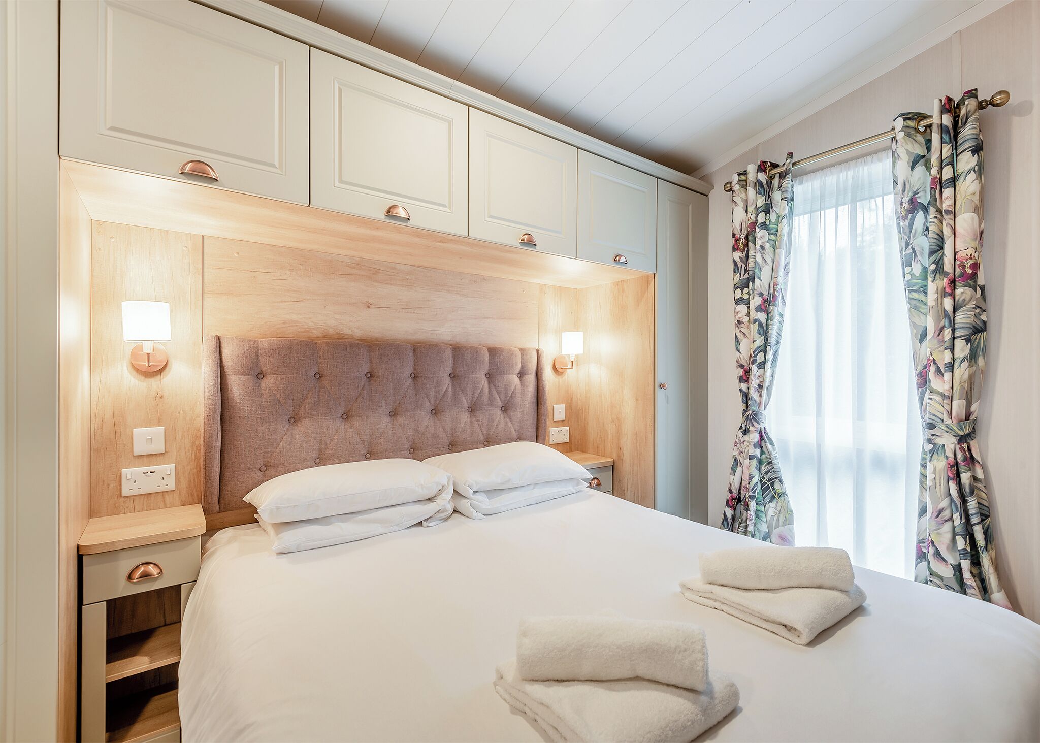 Quarto de hotel com uma cama grande, cabeceira de madeira, mesas de cabeceira e uma janela com cortinas florais.