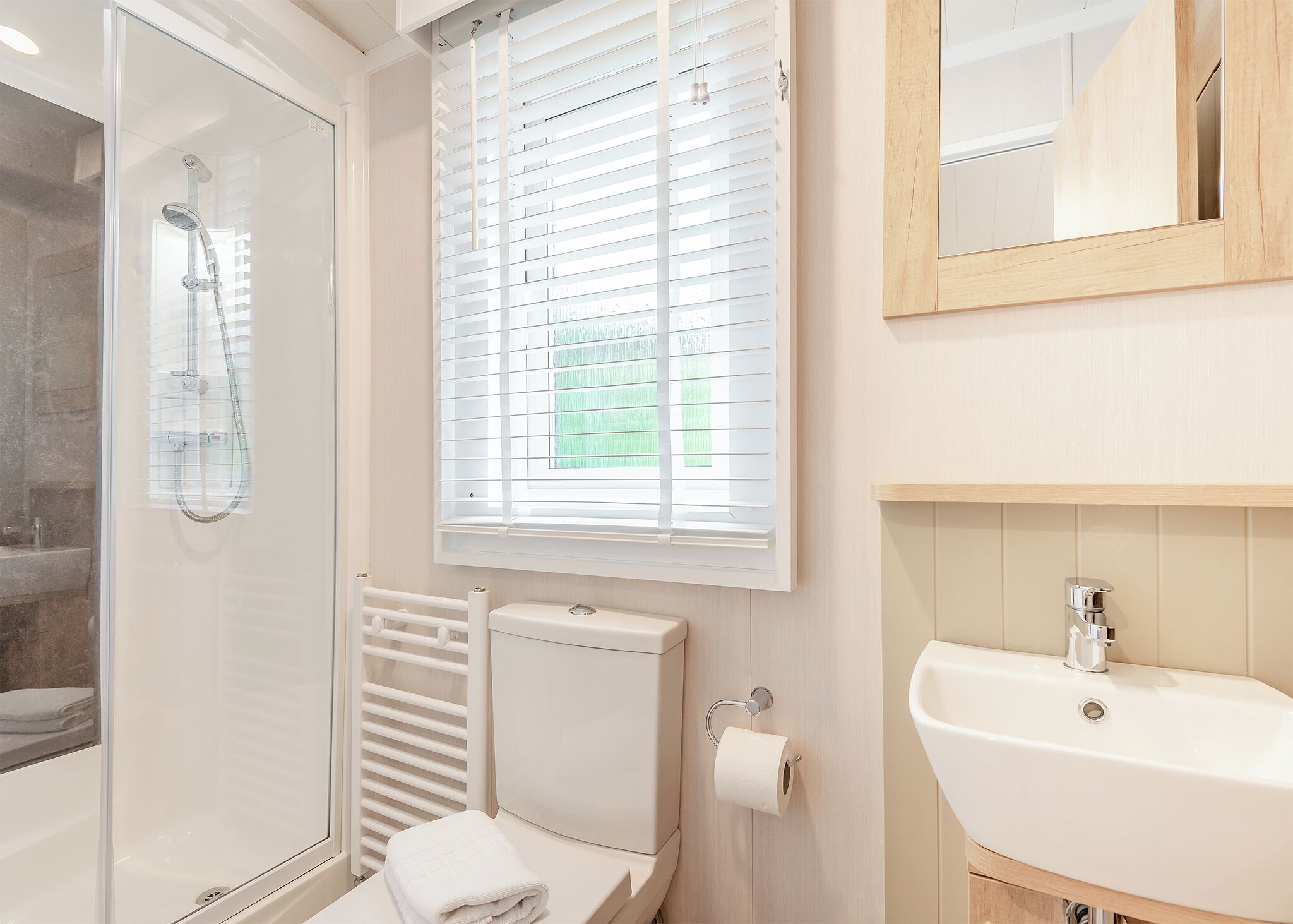 Un baño con cabina de ducha de vidrio, inodoro con soporte para toallas de papel, lavamanos y ventana con persianas.