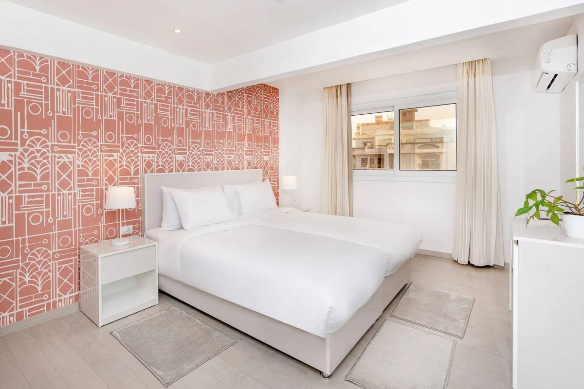 A modern hotel room with a large bed, a nightstand, a potted plant, and a window with curtains.