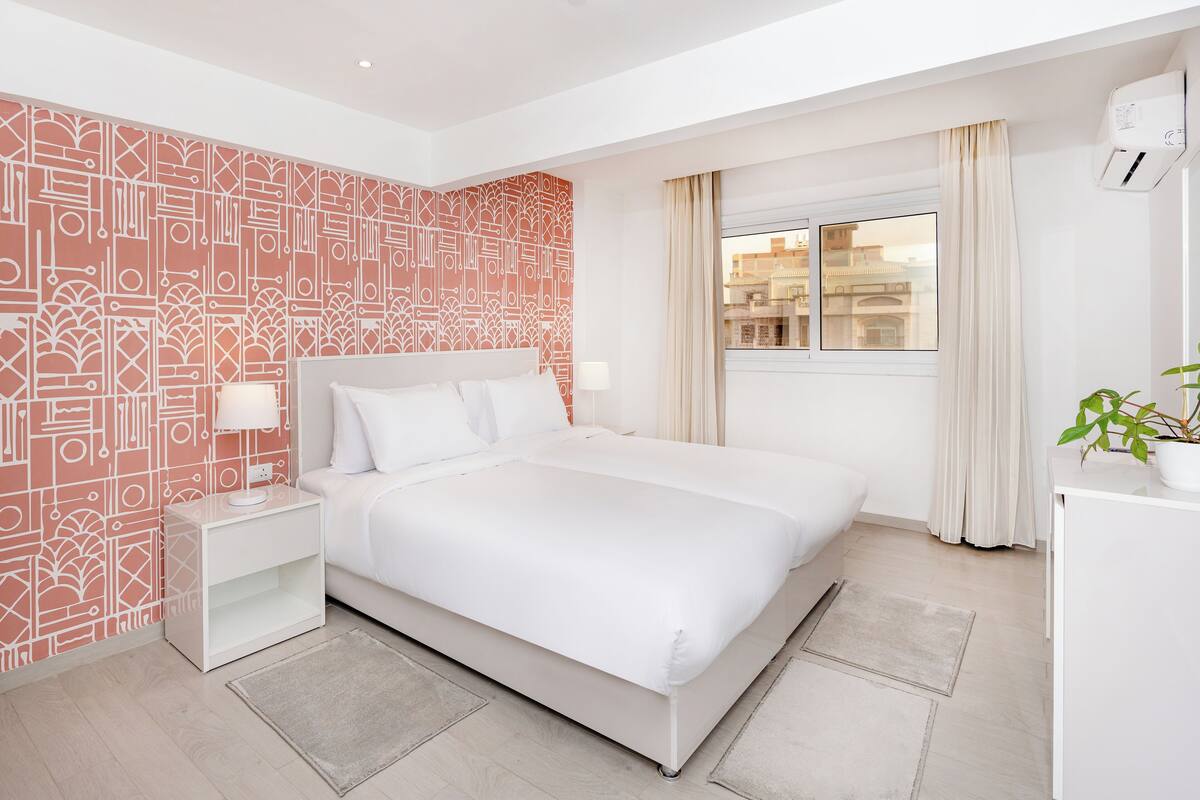 A modern hotel room with a large bed, a nightstand, a potted plant, and a window with curtains.