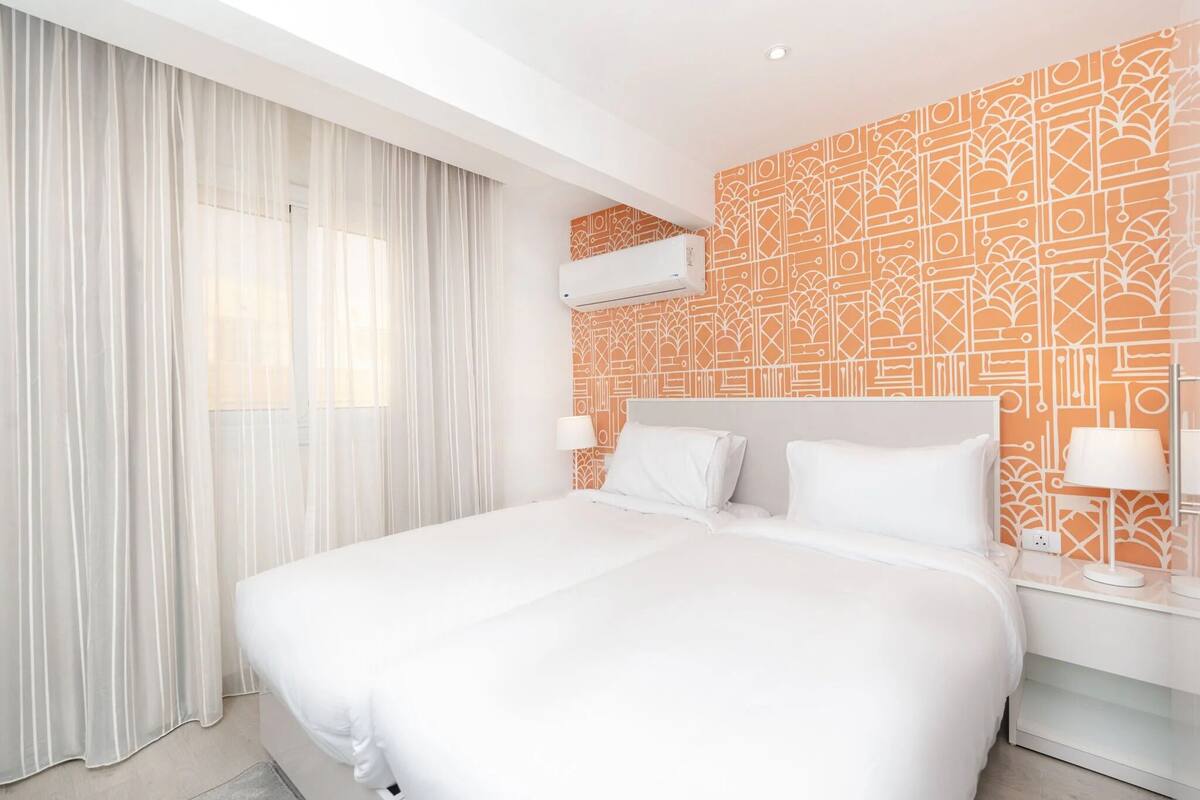 A hotel room with a large bed, white curtains, an air conditioning unit, and patterned wallpaper.