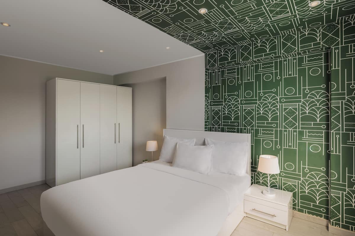 A bedroom with a large bed, white nightstands, a wardrobe, and a patterned wallpaper.