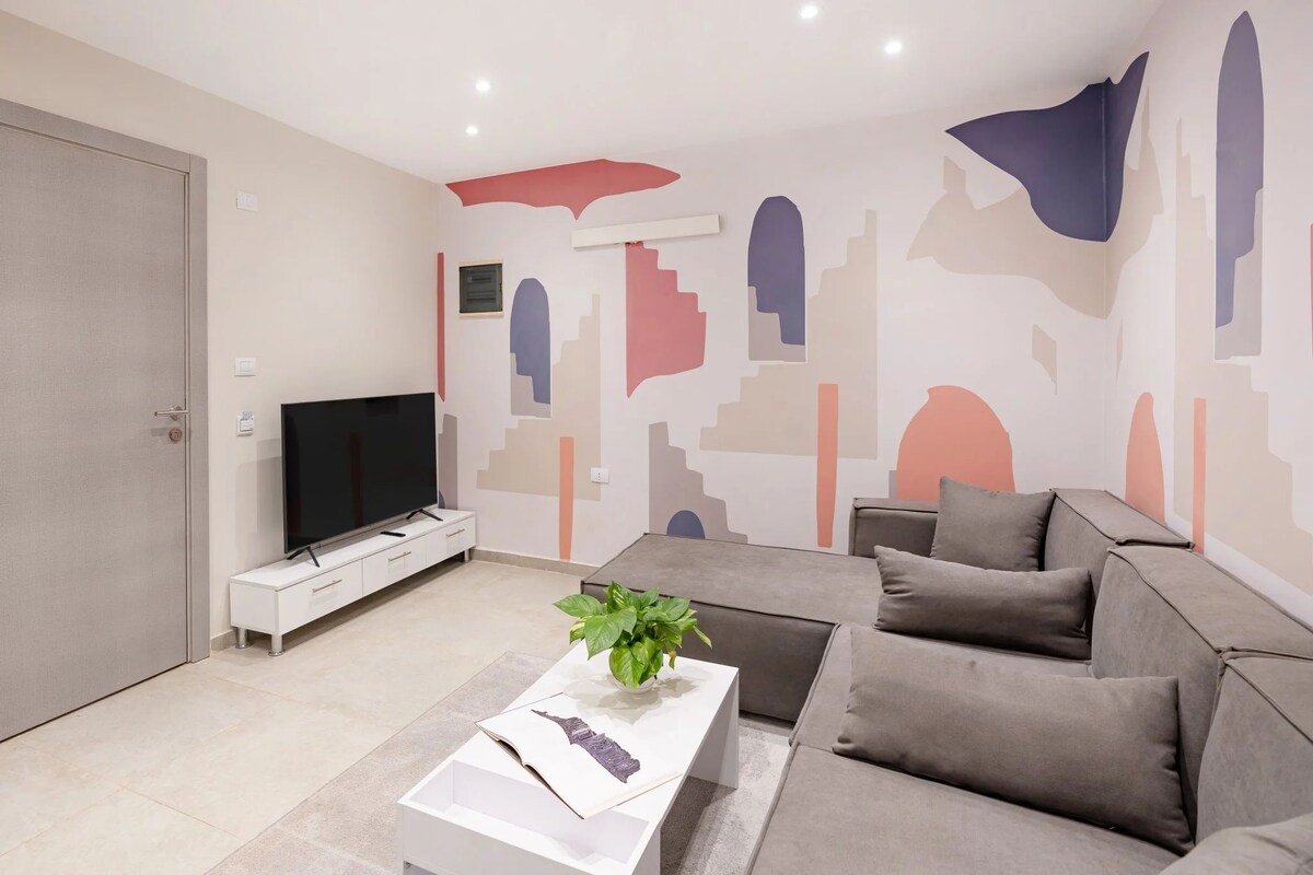 Ein modernes Wohnzimmer mit Fernseher, einer weißen Ablagetisch, einem grauen Sofa und abstrakter Wandkunst.