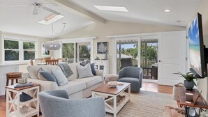 Ein geräumiges Wohnzimmer mit hoher Decke, Deckenventilator, großem Fenster, einer Couch, einem Couchtisch, einem Sessel und einem Fernseher.