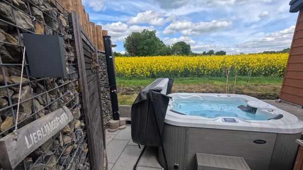 Et spabad med en træterrasse, en stenvæg og en blomstereng med gule blomster i baggrunden.