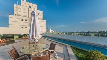 Uma área de piscina no terraço, com mesa de vidro, cadeiras de vime, um guarda-sol branco e vista para o horizonte da cidade.