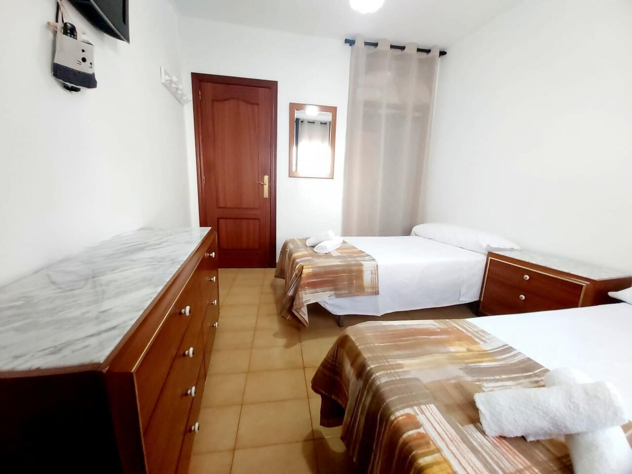 Habitación de hotel con tocador de madera, dos camas con ropa de cama a rayas, una puerta de madera, un espejo y una ventana con cortinas traslúcidas.