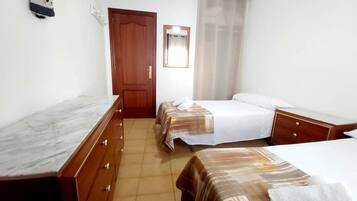 Habitación de hotel con tocador de madera, dos camas con ropa de cama a rayas, una puerta de madera, un espejo y una ventana con cortinas traslúcidas.