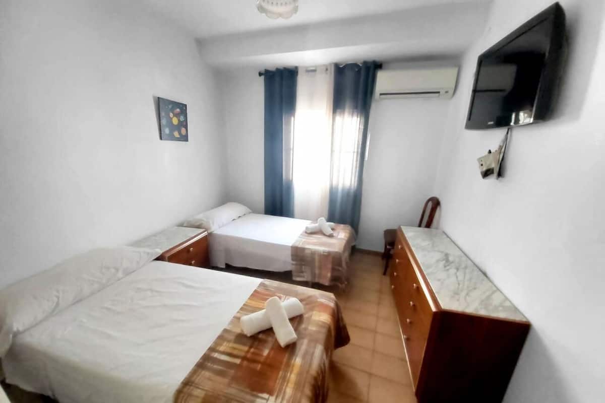 A hotel room with two beds, a wooden dresser, a TV mounted on the wall, and a window with curtains.