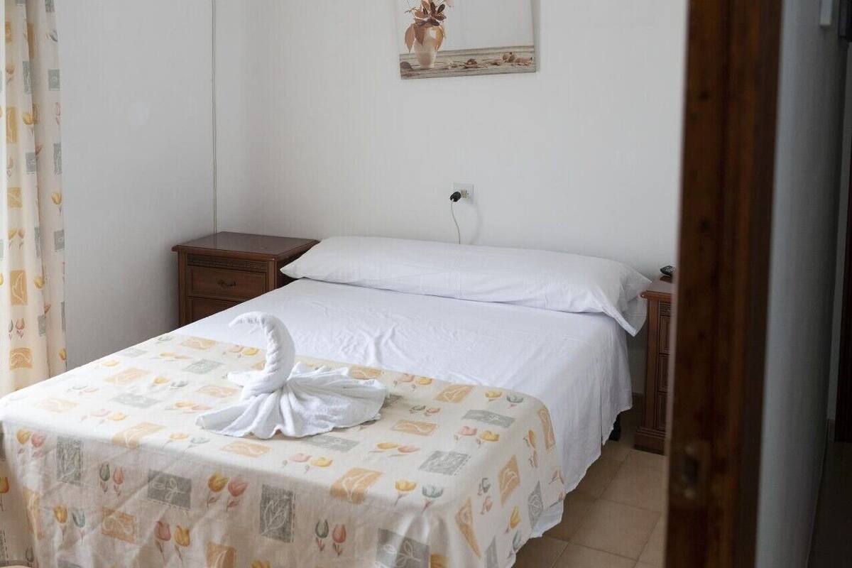 A neatly made bed with a white duvet and a decorative towel, a small wooden nightstand, and a wall-mounted power outlet.