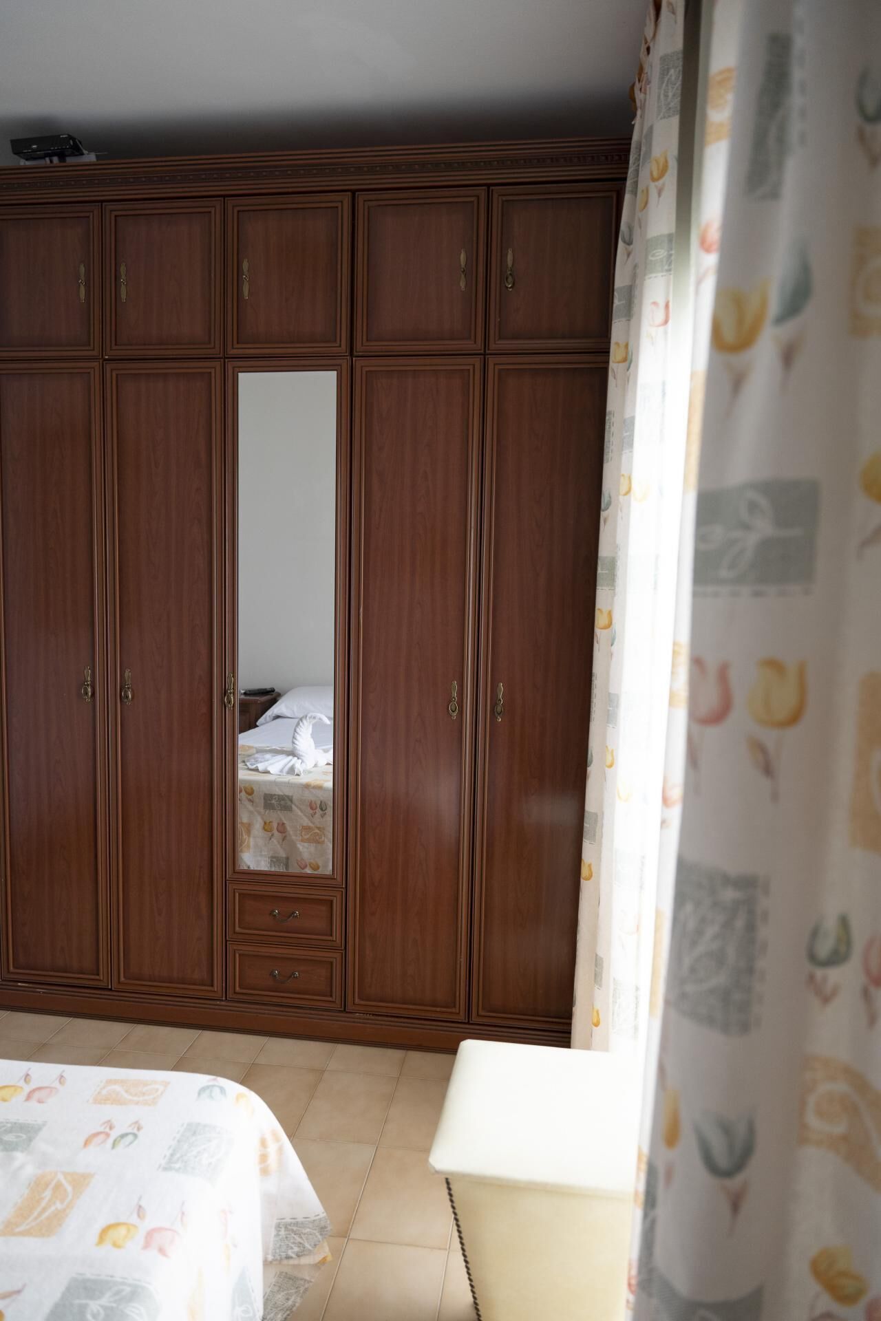 A bedroom with a large wooden wardrobe, a bed with a floral bedspread, and a bedside table.
