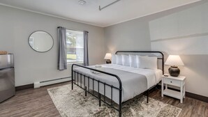 Een moderne slaapkamer met een metalen bed, een ronde spiegel, een nachtkastje met een lamp, en uitzicht op het landschap door een raam met gordijnen.