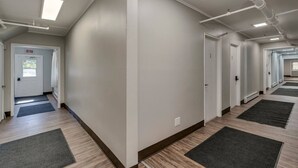 A clean, well-lit hallway with wooden flooring, dark mats at the entrance of each room, and white walls. - The Pine Street Inn - Room 4 (Watertown)
