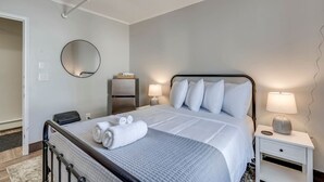 A neatly made bed with white linens and pillows, a bedside table with a lamp, a small refrigerator, and a round mirror on the wall. - The Pine Street Inn - Room 4 (Watertown)