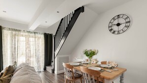 A dining area with a wooden table set for four, a black wall clock, and a staircase leading to an upper level. - Period Cottage Settle (Settle)