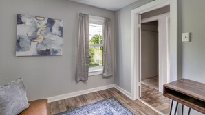 Une pièce avec une fenêtre, un tableau, un plancher en bois, un banc et une porte qui donne sur une autre pièce.