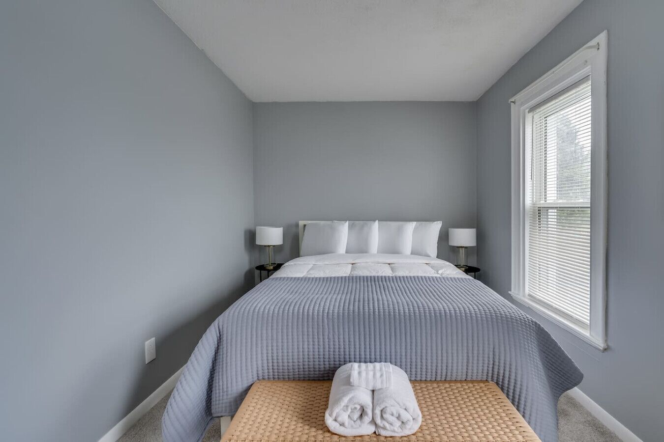 A bedroom with a bed, two bedside lamps, a window with blinds, and a small bench with towels.