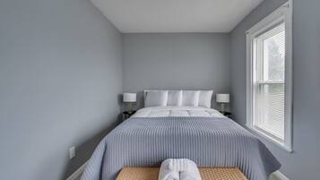 A bedroom with a bed, two bedside lamps, a window with blinds, and a small bench with towels.