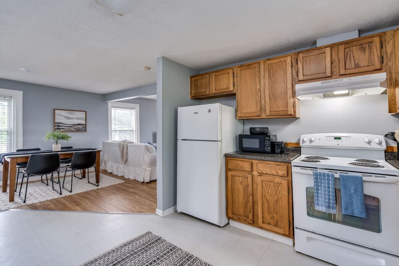 A modern kitchen with wooden cabinets, a white refrigerator, a microwave, a stove, and a dining area with a table and chairs.