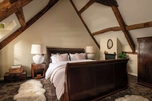 A bedroom with a sloped ceiling, a wooden bed, two bedside tables with lamps, a dresser, and a small plant. - Homestead - Cosy Cotswolds Cottage with Hot Tub (South Cerney)