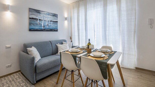 Un comedor con una mesa preparada para cuatro personas, un sofá gris y un cuadro de veleros en la pared.
