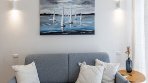 Una sala de estar moderna con un sofá gris, cojines blancos, un cuadro de veleros y un jarrón sobre una mesita auxiliar.