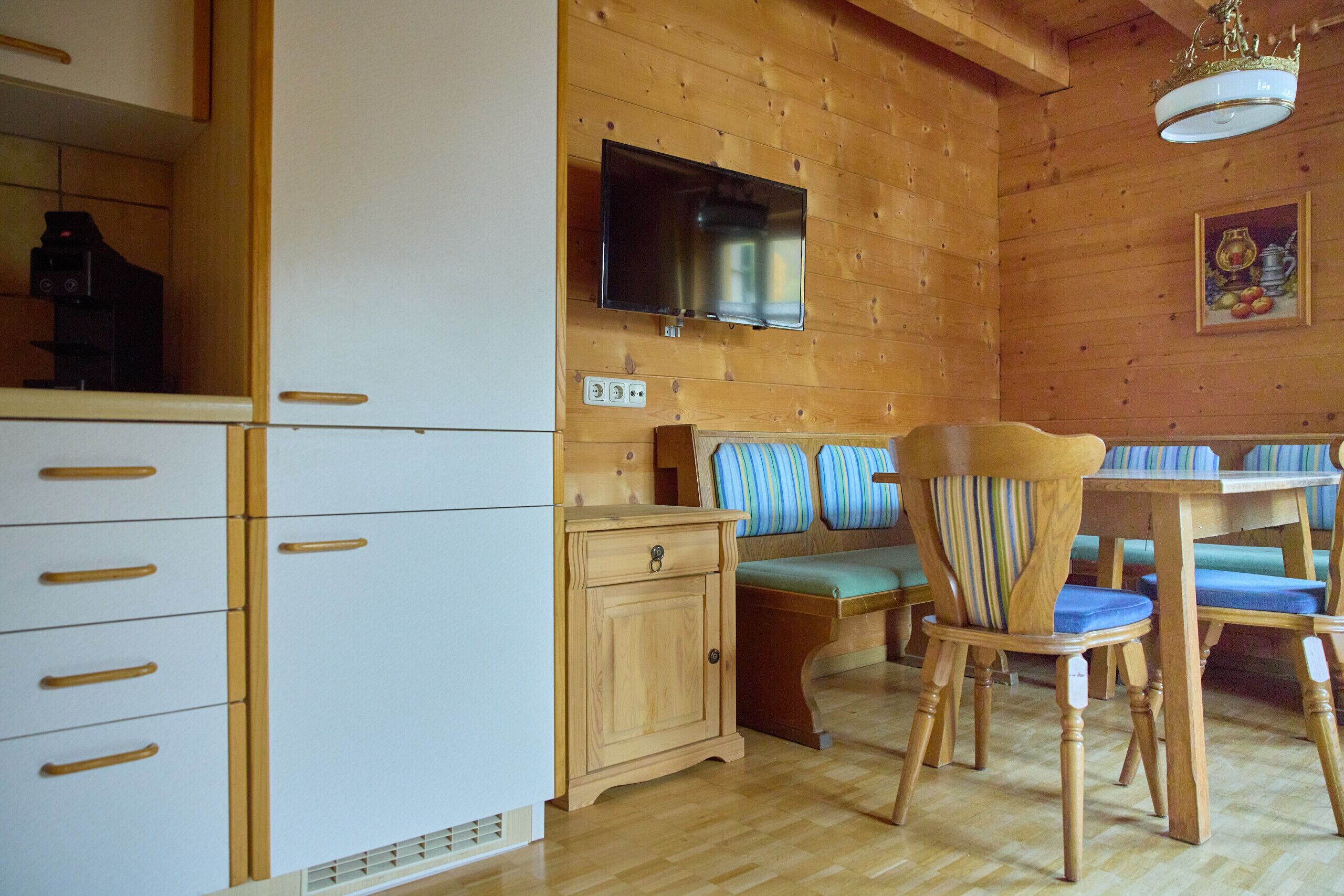 A wooden-paneled room with a television mounted on the wall, a wooden cabinet, a sofa with striped cushions, and a dining table with chairs.
