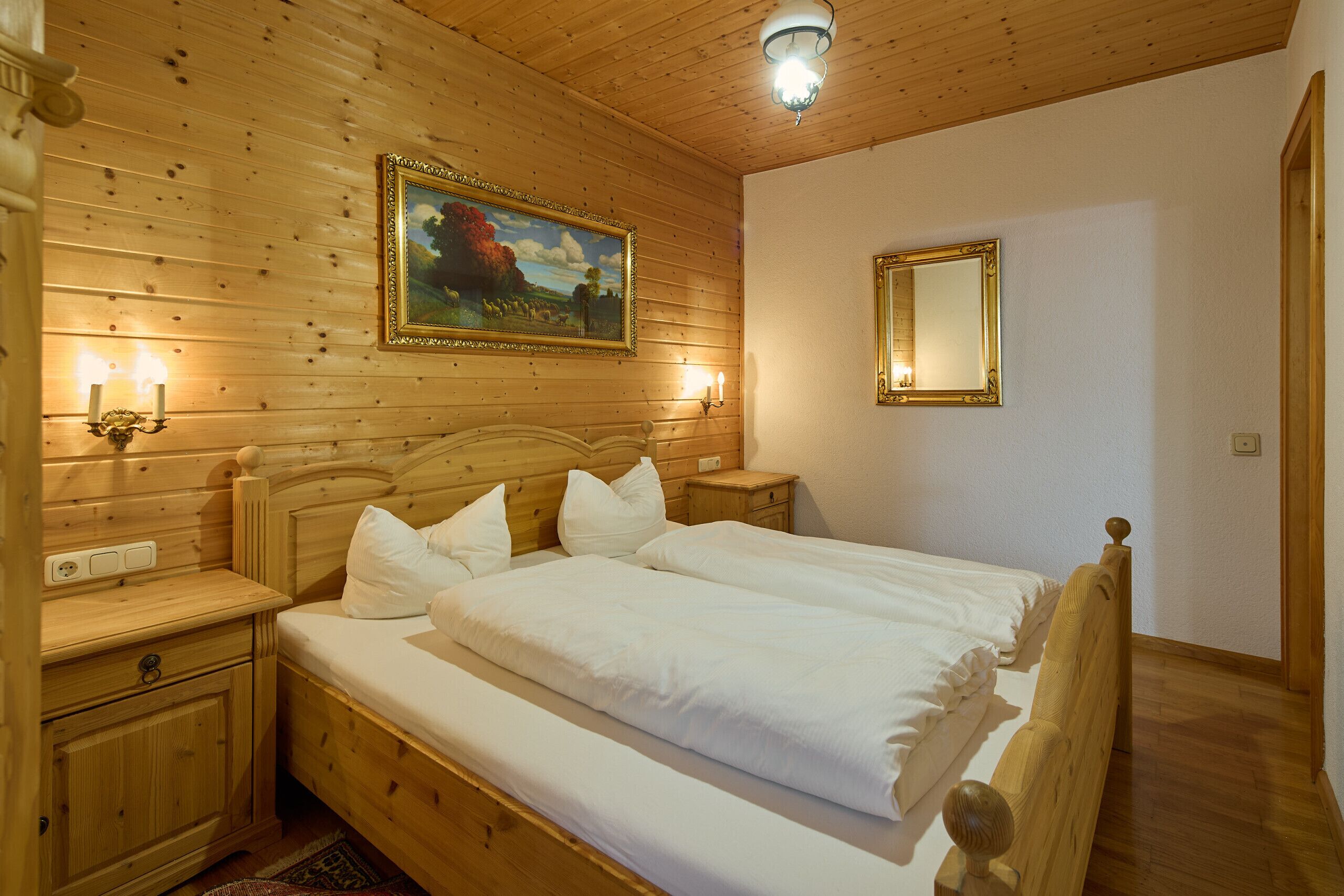 A wooden-paneled bedroom with a double bed, bedside lamps, a framed painting, and a mirror.