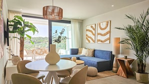 Ein modernes Wohnzimmer mit einem blauen Sofa, einem runden Esstisch und Stühlen. Zwei große Fenster mit Vorhängen bieten einen Blick ins Grüne, und an den Wänden befinden sich dekorative Kunstwerke.