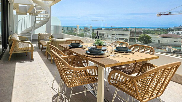 Eine Dachterrasse mit einem Holztisch, gedecktem Esstisch, Korbstühlen und Blick auf Stadt und Meer.