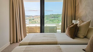 Ein Hotelzimmer mit zwei Betten, beiger Bettwäsche, Blick auf eine grüne Landschaft und einem Balkon mit Geländer.