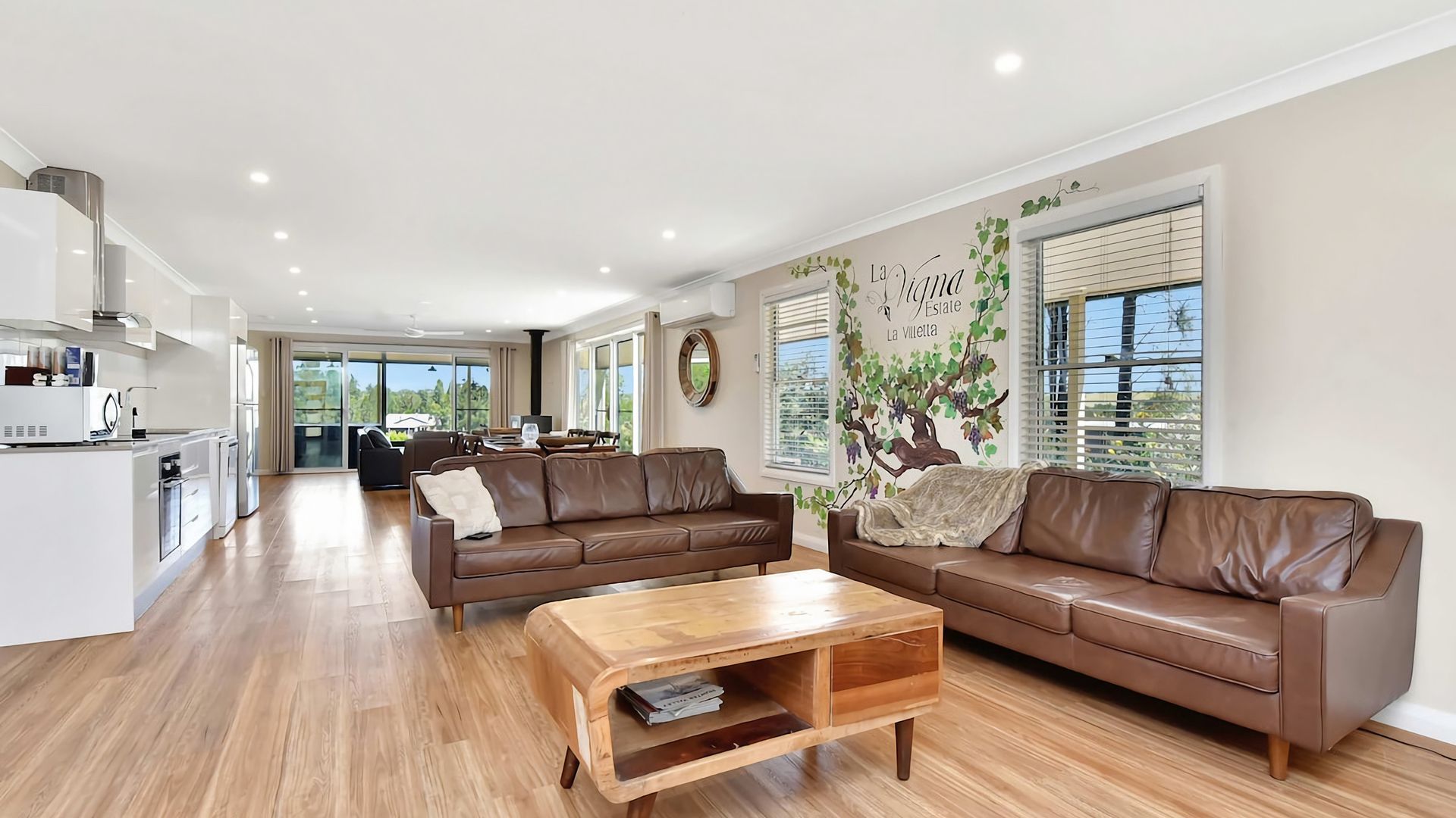 A spacious living area with a brown leather sofa, a wooden coffee table, and a kitchen in the background.