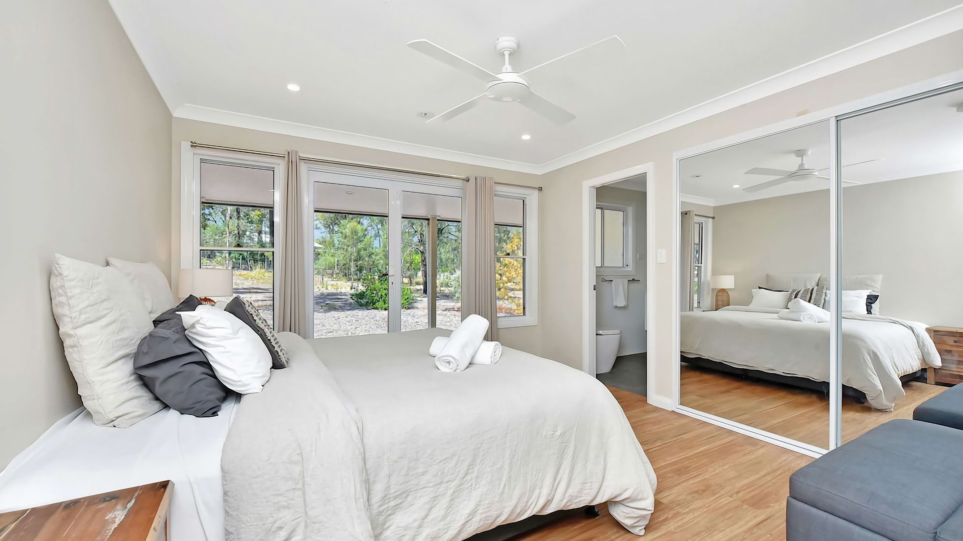 A bedroom with a bed, a ceiling fan, a large mirror, and a view of the outdoors.