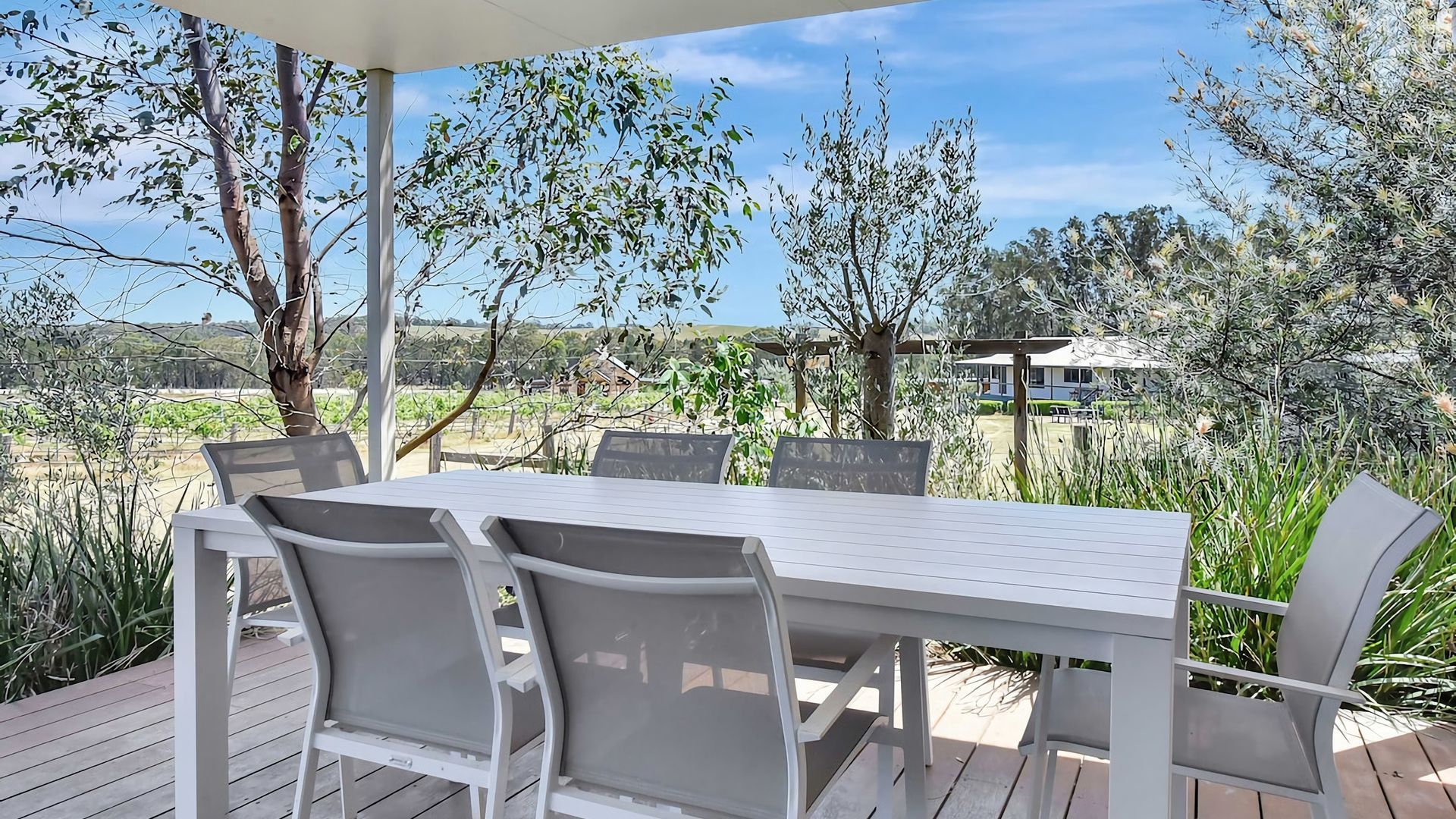En udendørs terrasse med et hvidt bord og stole, omgivet af grøn vegetation og træer.