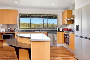 A modern kitchen with wooden cabinets, stainless steel appliances, and a large window offering a view of a forest. - Shiraz Homestead (Pokolbin)