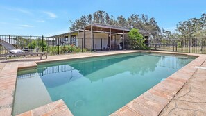 A rectangular swimming pool with a stone border, a covered patio area, and a fenced yard with lounge chairs.