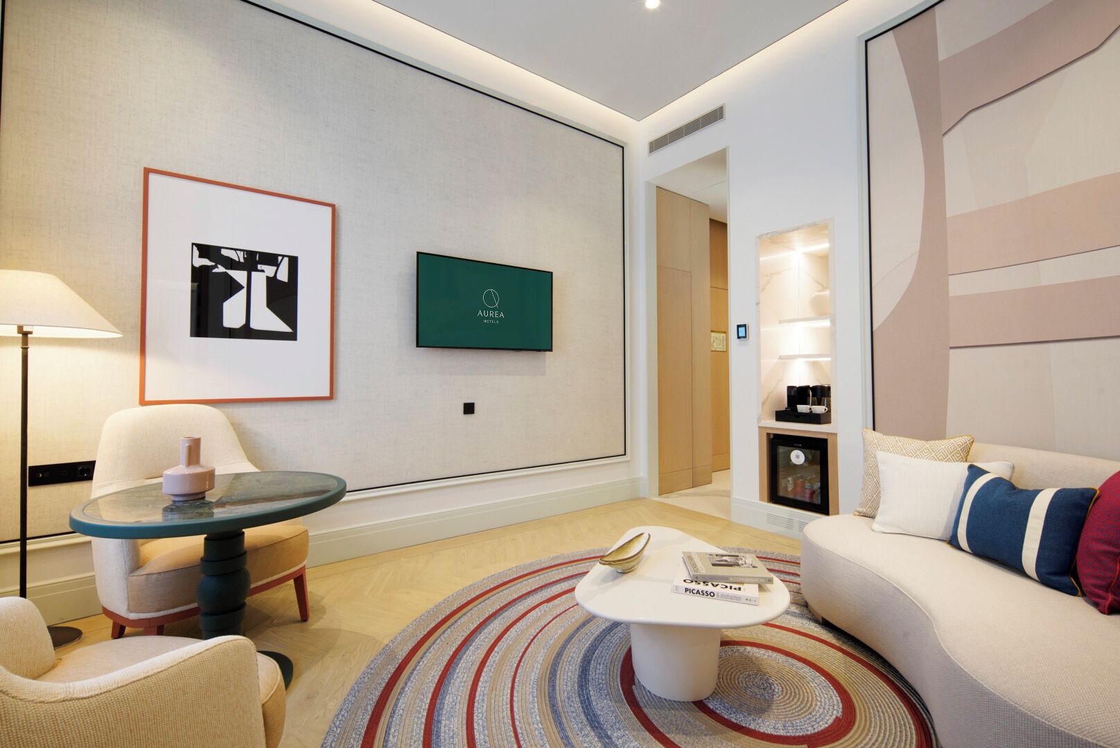 Una habitación de hotel moderna con una mesa de centro redonda, un sofá, una silla, una lámpara y obras de arte en las paredes.