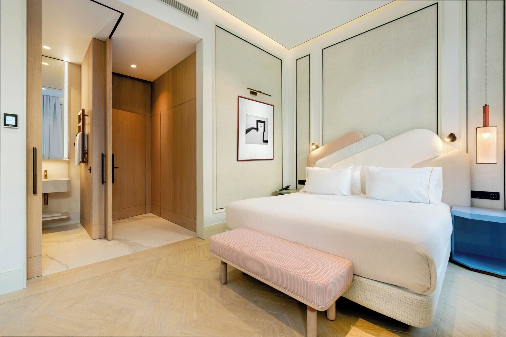 Una habitación de hotel moderna con una cama grande, un banco, un vestidor y un cuadro en la pared.