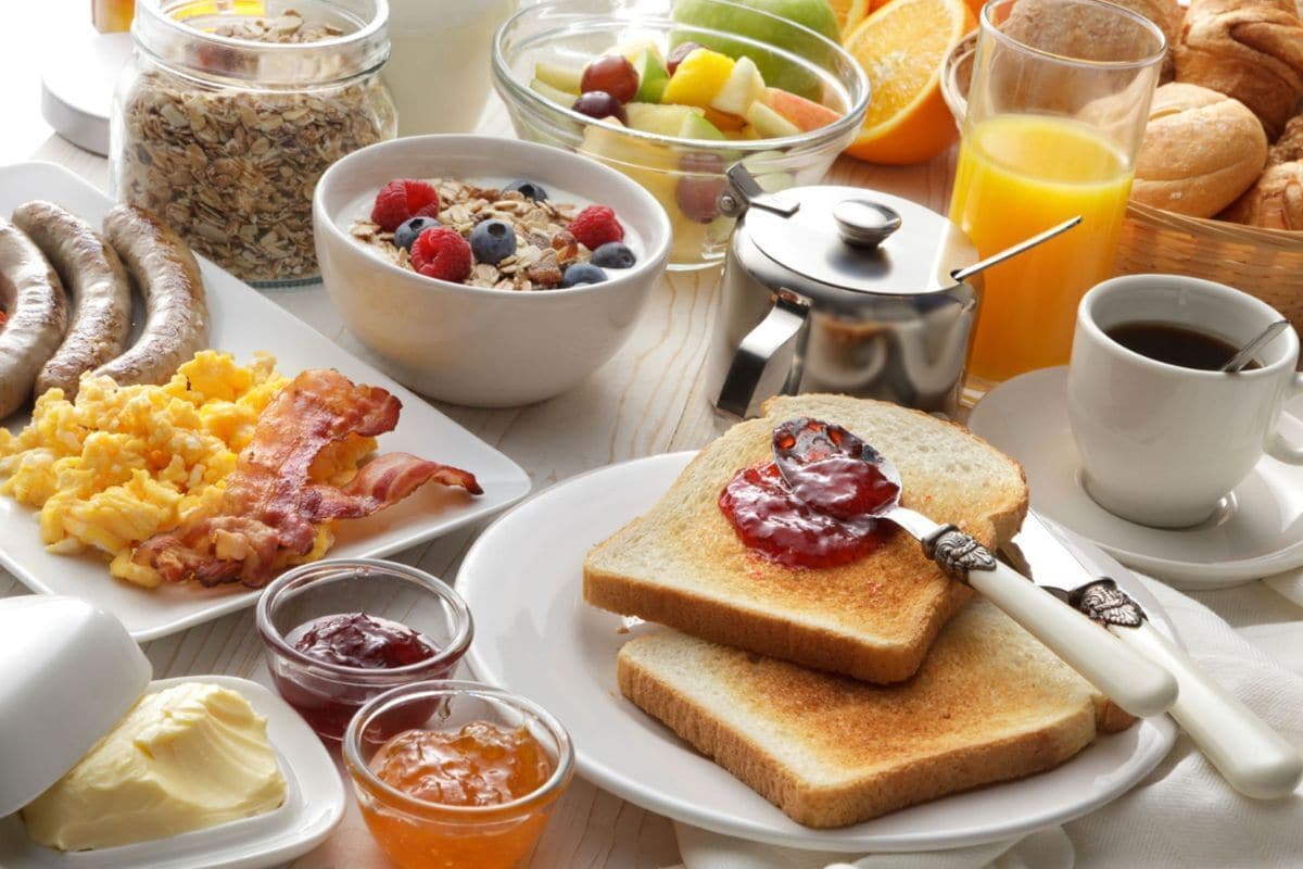 Una mesa de desayuno con diversos alimentos, como tostadas con mermelada, huevos revueltos, salchichas y un bol de fruta.