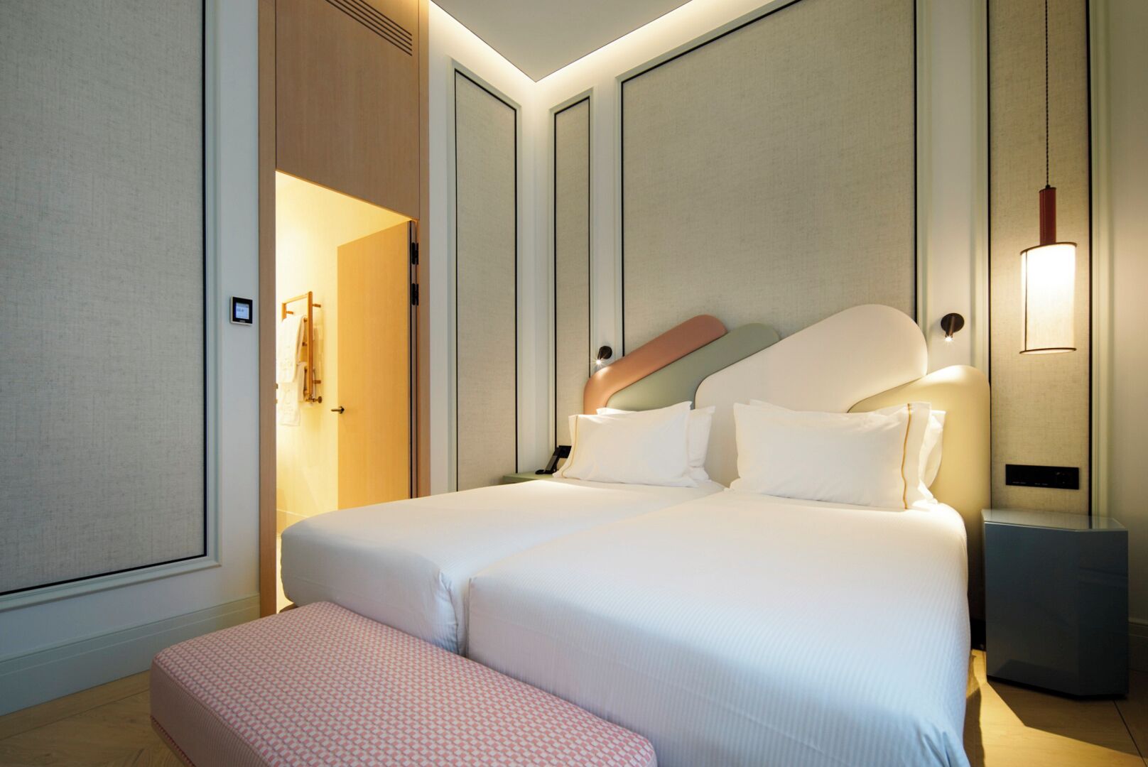 Una cama doble con cabecera, mesitas de noche, un banco e iluminación de pared.