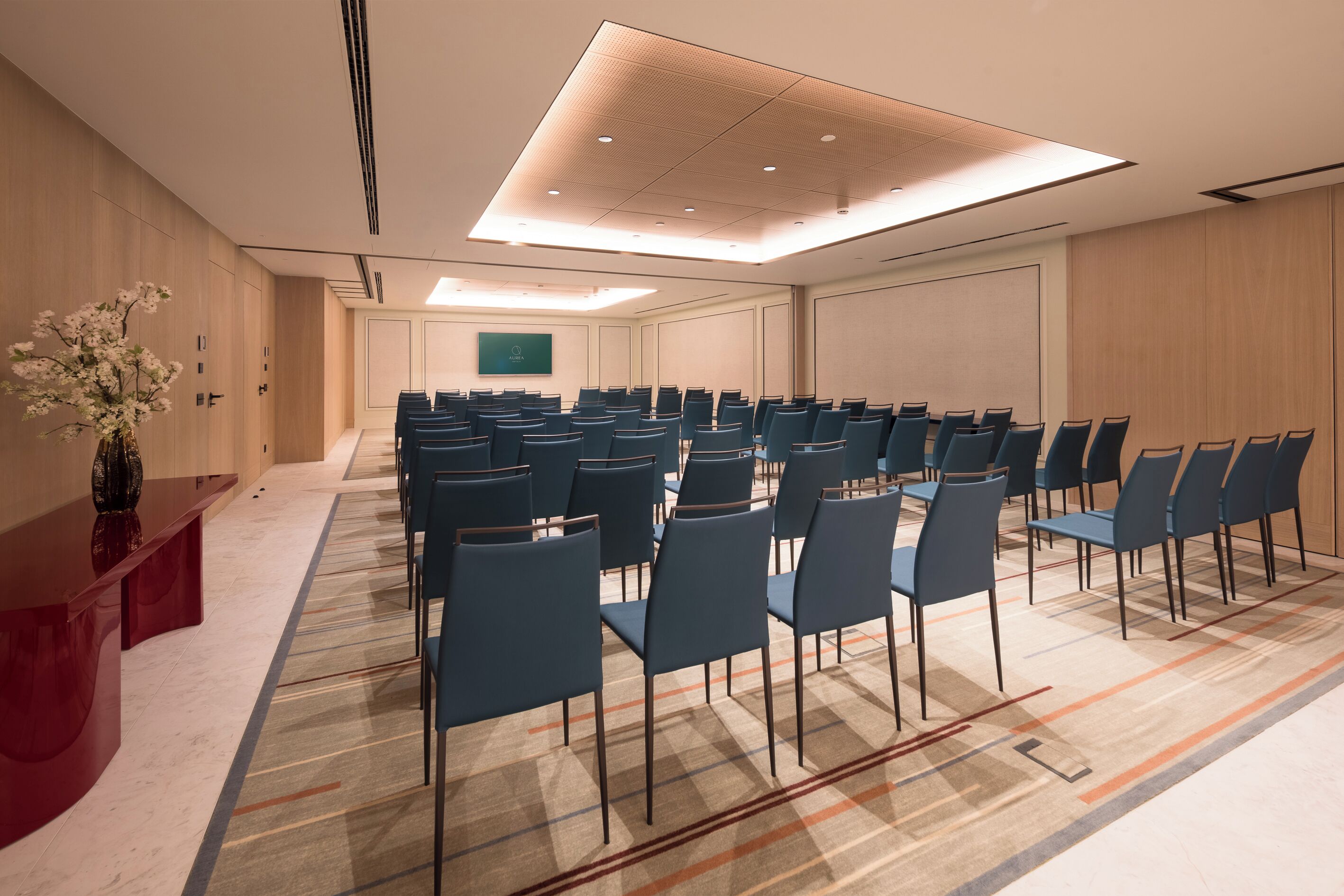 Un salón de conferencias con filas de sillas, un mostrador de recepción rojo y una pantalla grande en la pared.