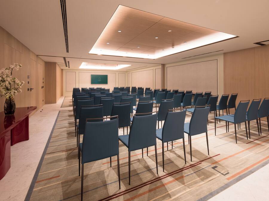 Un salón de conferencias con filas de sillas, un mostrador de recepción rojo y una pantalla grande en la pared.