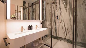 Un baño moderno con lavamanos de mármol blanco, dos grifos y una ducha con cerramiento de vidrio.