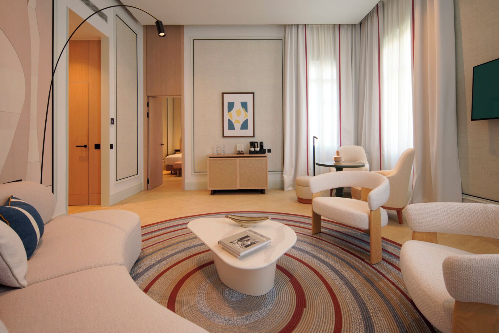 A modern living room with a sofa, armchairs, a coffee table, and a side table. There is a door leading to another room, a small cabinet, and a framed picture on the wall.